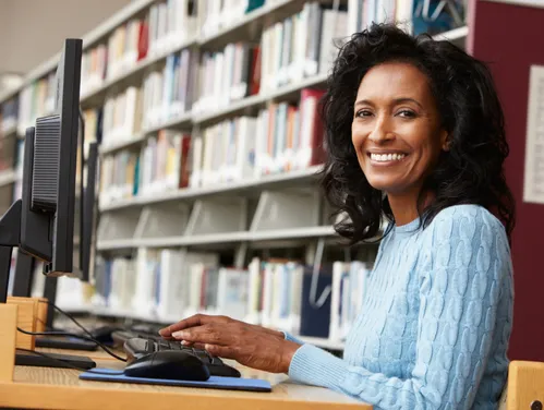 Woman using a computer in a library, she is looking at the camera and smiling.