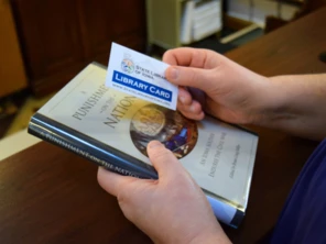 Closeup of hands holding a State Library Card and a book to check out.