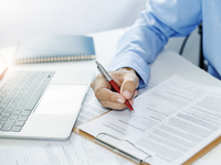 Closeup of a desktop with person's hand completing paperwork on a clipboard next to an open laptop.
