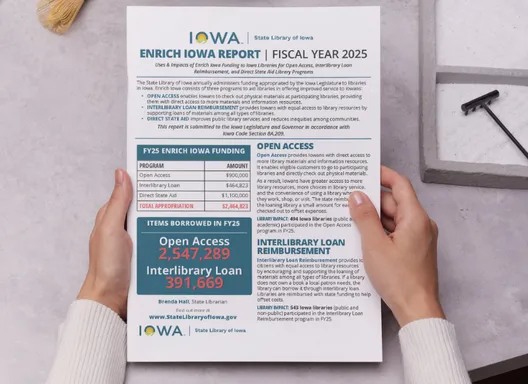 Hands on a tabletop holding a print copy of the Enrich Iowa Legislative Report.