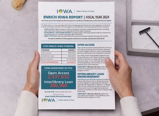Hands on a tabletop holding a print copy of the Enrich Iowa Legislative Report.
