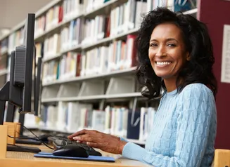 Woman using a computer in a library, she is looking at the camera and smiling.
